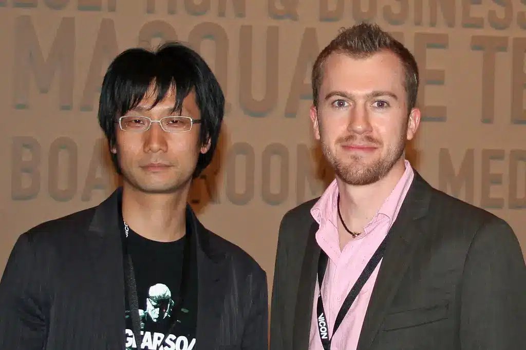 Hideo Kojima يرتدي نظارات وسترة سوداء، ويقف بجانب رجل آخر.